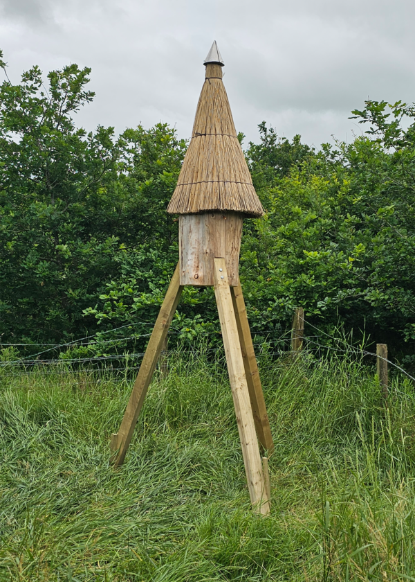 Elevated log hive in a field with fencing and trees behind